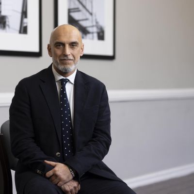 Khalid Naseem in a dark suit sitting in front of two pictures.