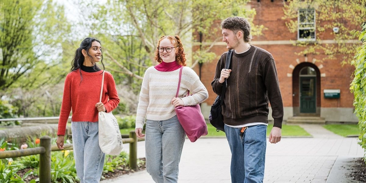 Three students walking on campus, smiling and chatting. There are flowers and trees and a red brick building in the background