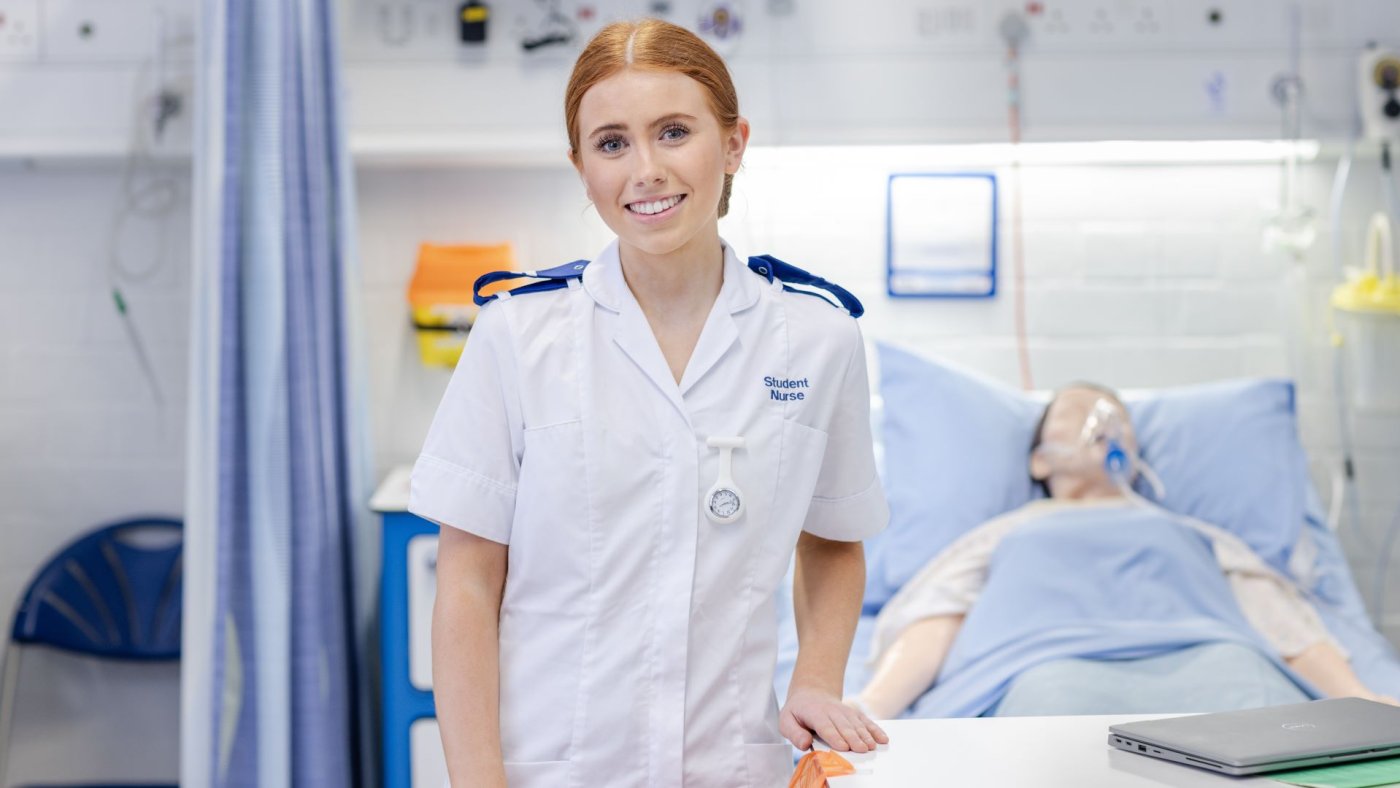 An adult nursing students stands smiling at the camera in the School of Healthcare clinical skills suite