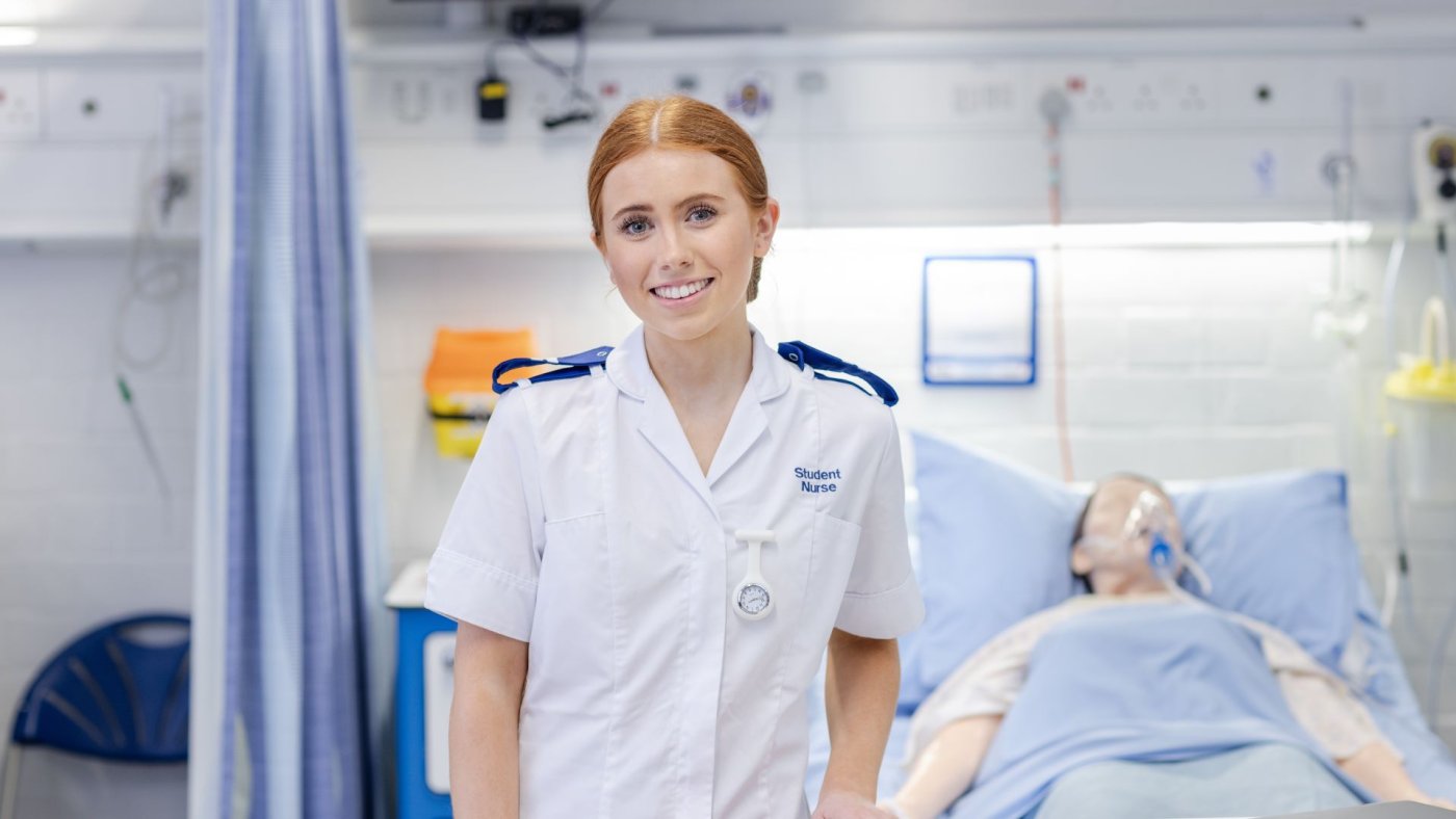 An Adult nursing student stands smiling at the camera in the clinical skills suite