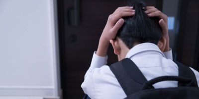 A child facing away, wearing a white school shirt and black backpack with their hands on their head.