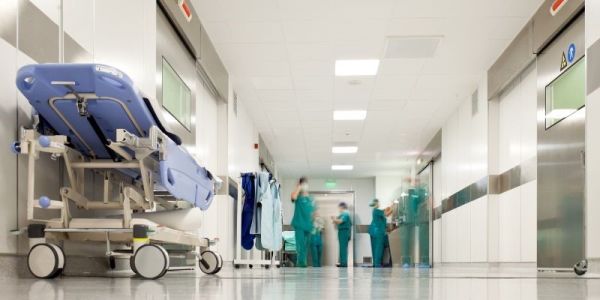 A hospital corridor with a bed to the left and staff members in scrubs at the far end.