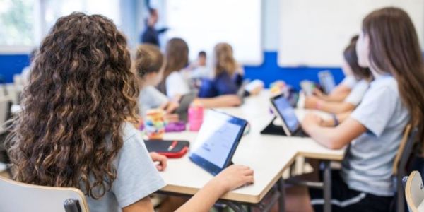 The back of two school children's heads. On the desk they have tablets and pencil cases. The teacher can be seen in the background.