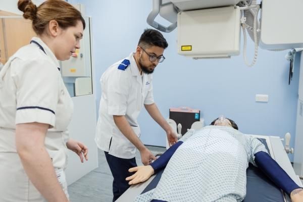 Teacher and male student examining dummy laid on table, both in clinical attire.