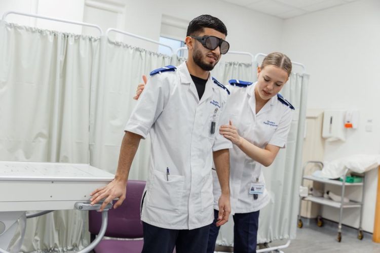 Female student supporting and guiding a male student wearing eye wear to a seat.