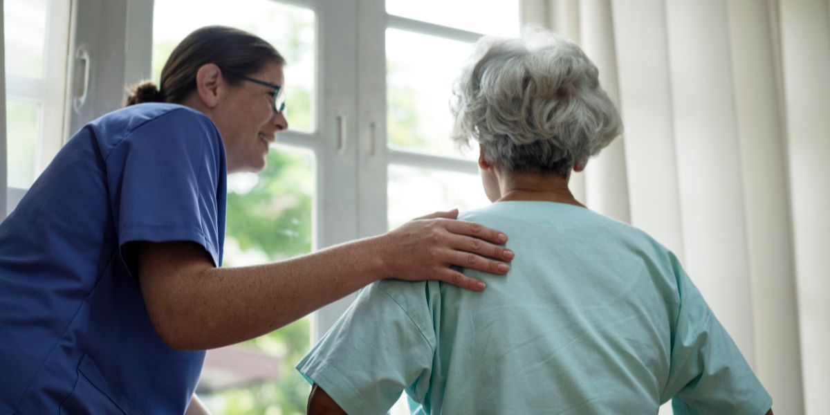A view from behind of a carer with their hand on the shoulder of an elderley patient in front of a large glass window.