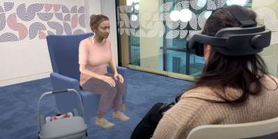 A student wearing a virtual reality headset, interacting with Stacey the virtual patient, who is sitting on a chair with her baby next to her in a baby carrier