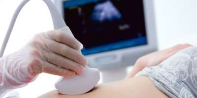An ultrasound device on a person's stomach. There is a screen in the background showing the ultrasound, it is blurred.