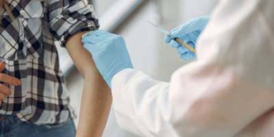 Close up of a medical professional in white coat and blue surgical gloves about to administer drugs into the upper arm of someone wearing a black and white checked shirt with their sleeve rolled up.